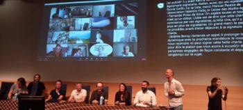Plusieurs personnes sont assises le long d'une table, elles sont souriantes, une autre personne est débout à côté d'une interprète en langue des signes. En arrière plan, sur un écran, la transcription simultanée des échanges et sur un autre écran, la galerie des participants à distance sur la visioconférence.